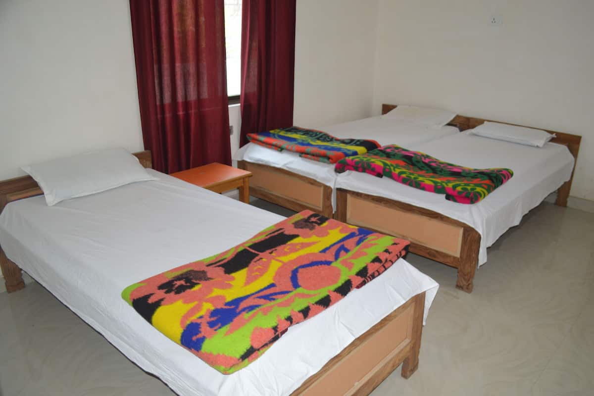 Quarto com três camas de solteiro, cada uma com estrutura de madeira e roupa de cama branca. Pequeno criado-mudo de madeira com abajur ao lado de cada cama. O quarto tem um design simples e funcional, com paredes lisas e chão de azulejos.