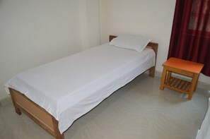 A single bed with a wooden frame and white bedding, a small wooden nightstand beside it, and a red curtain partially visible in the background. - Middle Way Home Stay (Gaya)