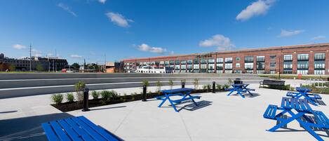 A spacious outdoor area with multiple blue picnic tables and benches, a large brick building in the background, and a clear blue sky.