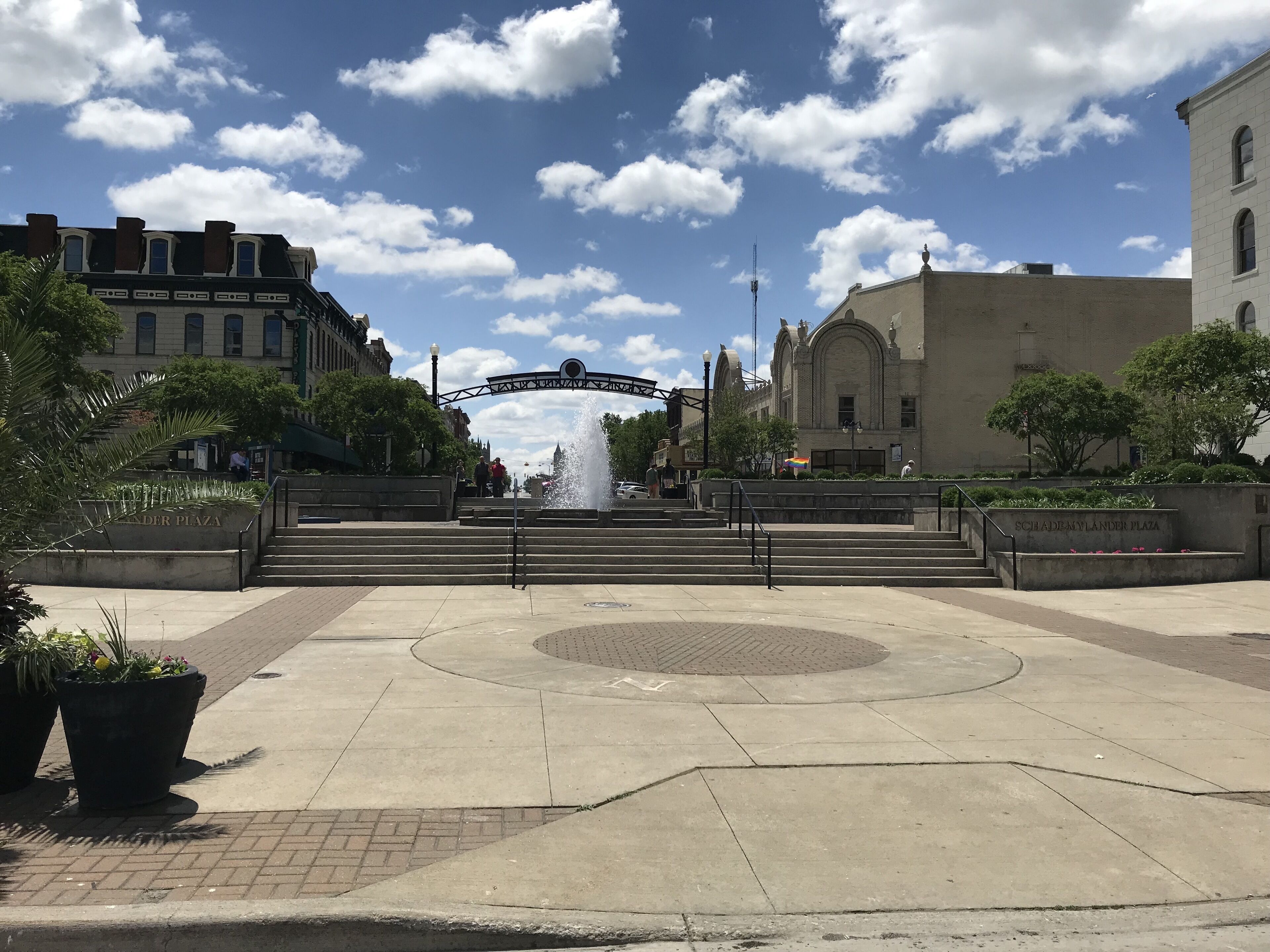 A public square with a fountain, steps leading to a building, and a clear sky.
