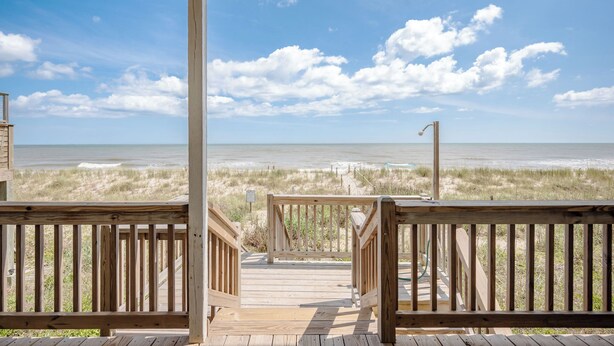 Eine Holzterrasse führt zu einem Blick auf den Strand bei blauem Himmel.