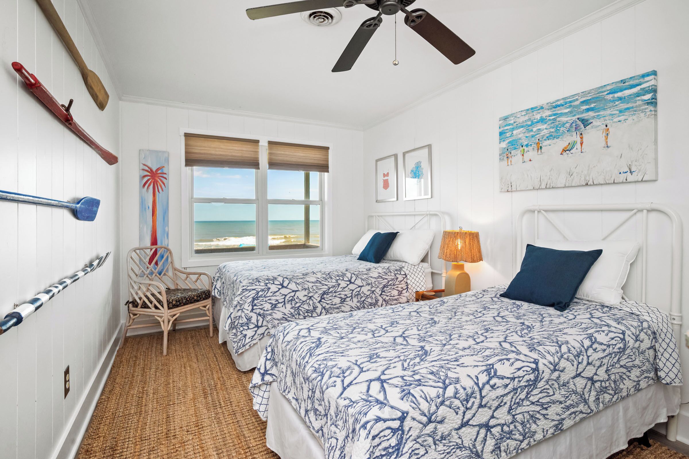 A room with two beds, a ceiling fan, a beach view, and a painting of a beach scene.