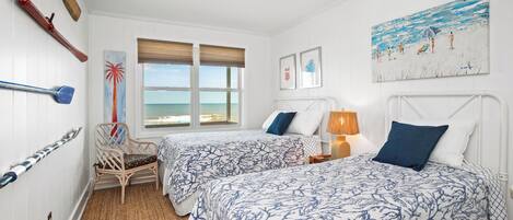 A room with two beds, a ceiling fan, a beach view, and a painting of a beach scene.