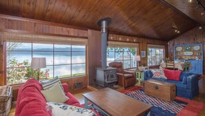 Een knus cabin interieur met een groot houten plafond, een houtkachel, een rode bank, een blauwe slaapbank, en uitzicht op een meer door grote ramen.