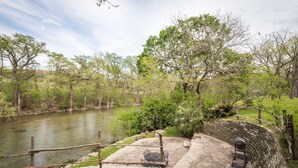 A patio with stone flooring, a grill, and a view of a river and trees. - Braun Rio - Guadalupe River, 2 King Master Suites, Loft, Riverfront Patio (New Braunfels)