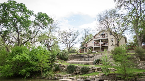 A stone building with a balcony overlooking a river, surrounded by lush greenery. - Braun Rio - Guadalupe River, 2 King Master Suites, Loft, Riverfront Patio (New Braunfels)