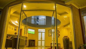Un hall de hotel con un separador de vidrio, una recepción, una chimenea, un televisor y un jarrón grande.