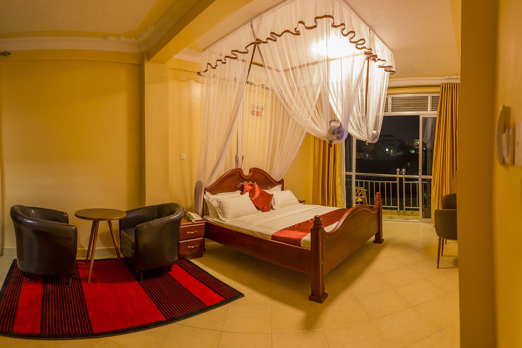 Een hotelkamer met een bed, nachtkastje, stoel, klein tafeltje, een rood en zwart vloerkleed, en een balkon met gordijnen.