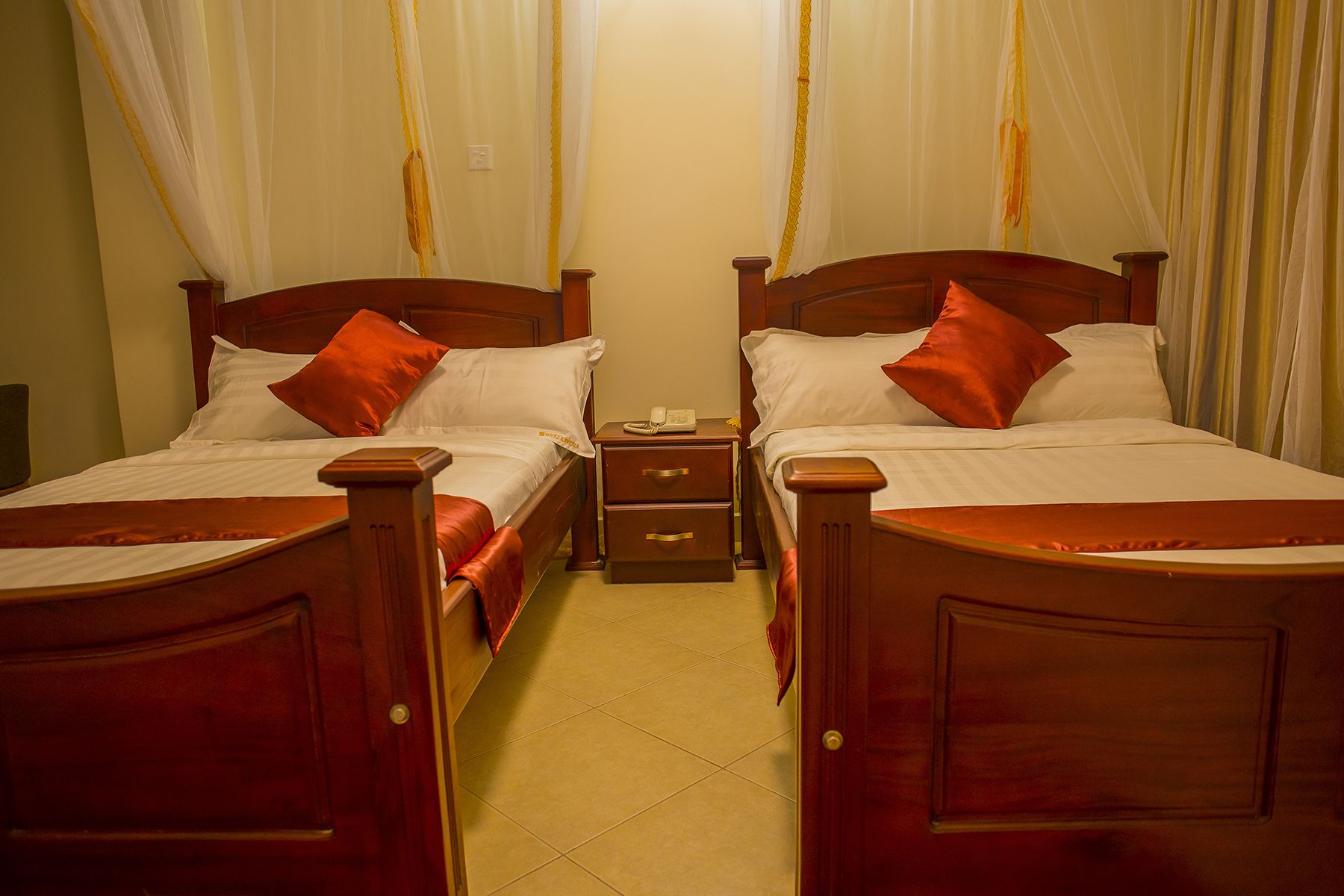 A hotel room with two wooden beds, each with white linens and orange pillows. There is a nightstand between the beds with a telephone and a small table. The room has a tiled floor and a curtain with a yellow trim.