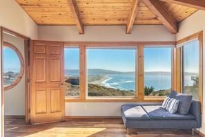 A room with a wooden ceiling, large windows with a coastal view, a wooden door, and a grey sofa with cushions. - Gorgeous 5 bedroom Home With Fabulous Ocean Views and 4 Master Suites (Dillon Beach)