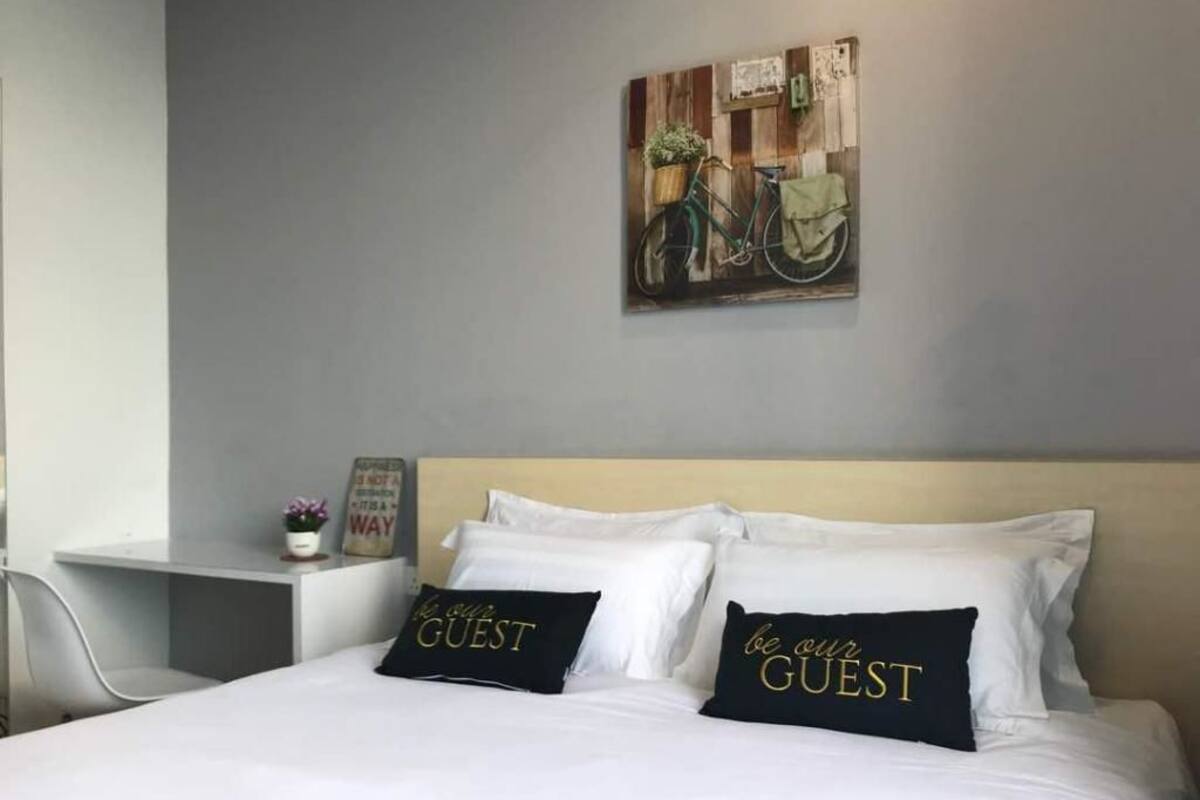Une chambre d’hôtel avec un lit, deux oreillers ornés de l’inscription « soyez les bienvenus », une petite table avec une plante et un tableau représentant un vélo, accroché au mur.