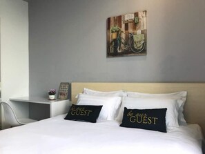 A hotel room with a bed, two pillows with 'be our guest' text, a small table with a plant, and a wall-mounted painting of a bicycle. - Symphony Suite Malacca (Malacca City)