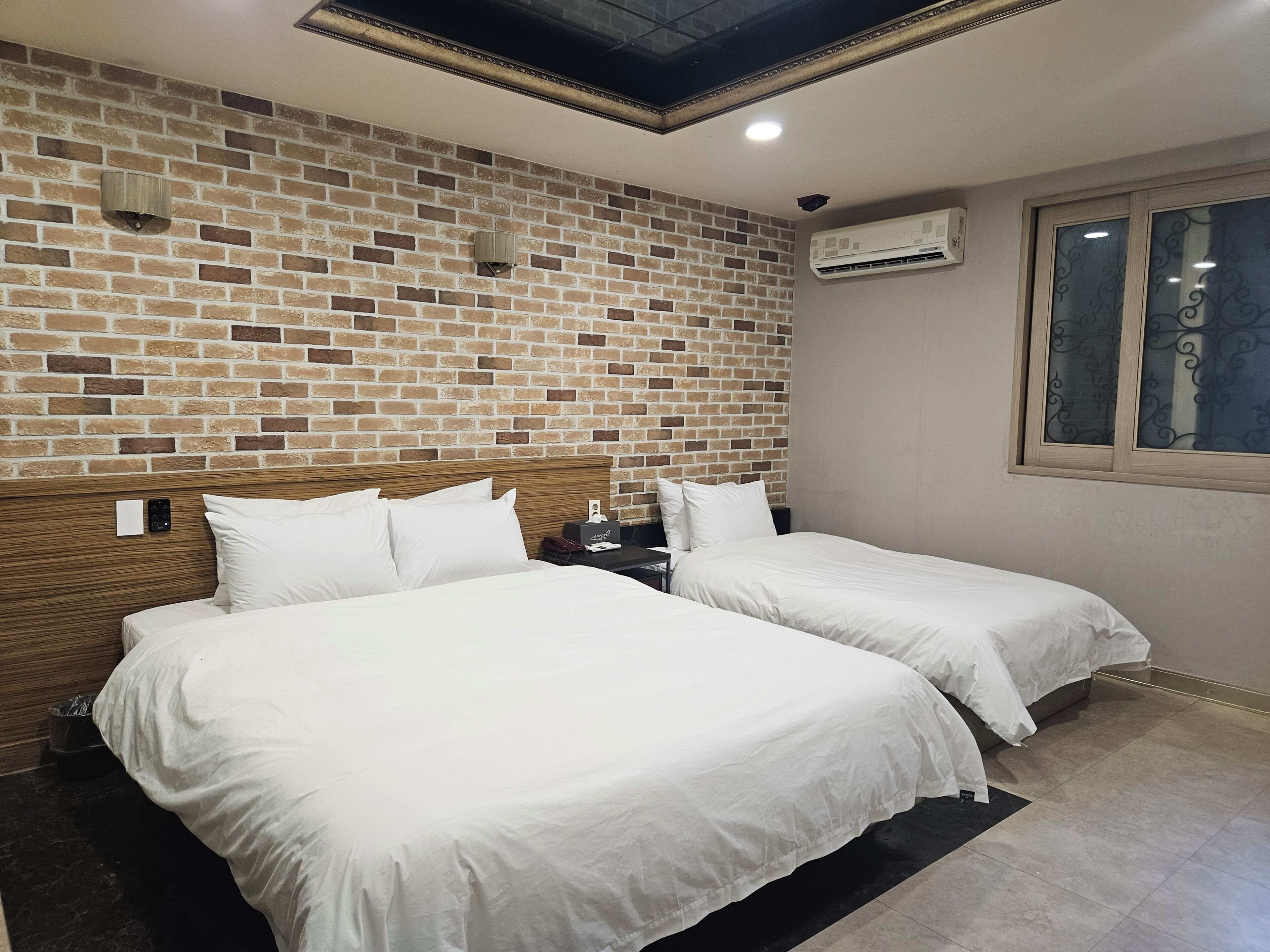 Une chambre d’hôtel avec un mur décoré d’un motif en briques, deux lits, une tête de lit en bois, un appareil de climatisation et une fenêtre ornée de vitraux.