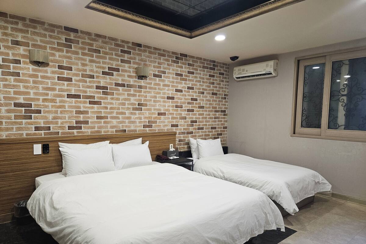 Une chambre d’hôtel avec un mur décoré d’un motif en briques, deux lits, une tête de lit en bois, un appareil de climatisation et une fenêtre ornée de vitraux.