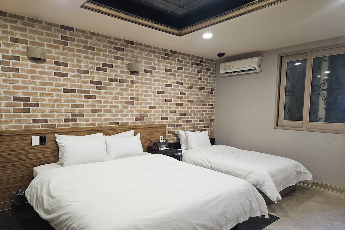Une chambre d’hôtel avec un mur décoré d’un motif en briques, deux lits, une tête de lit en bois, un appareil de climatisation et une fenêtre ornée de vitraux.