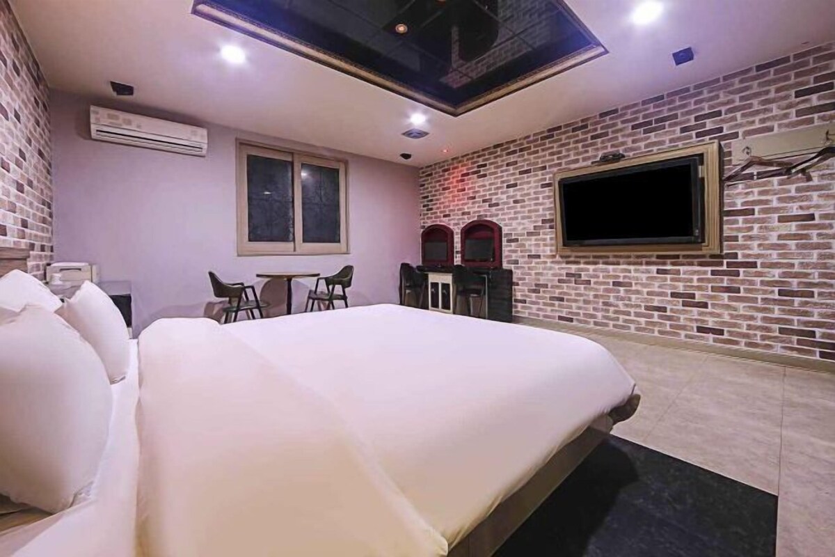 A hotel room with a large bed, a desk, two chairs, a TV, and a brick wall.