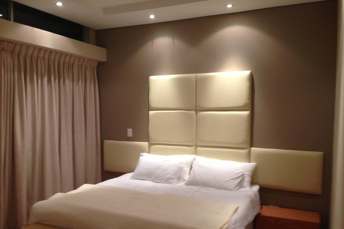 A modern hotel room with a large bed, a headboard with a light panel, and a wooden nightstand.