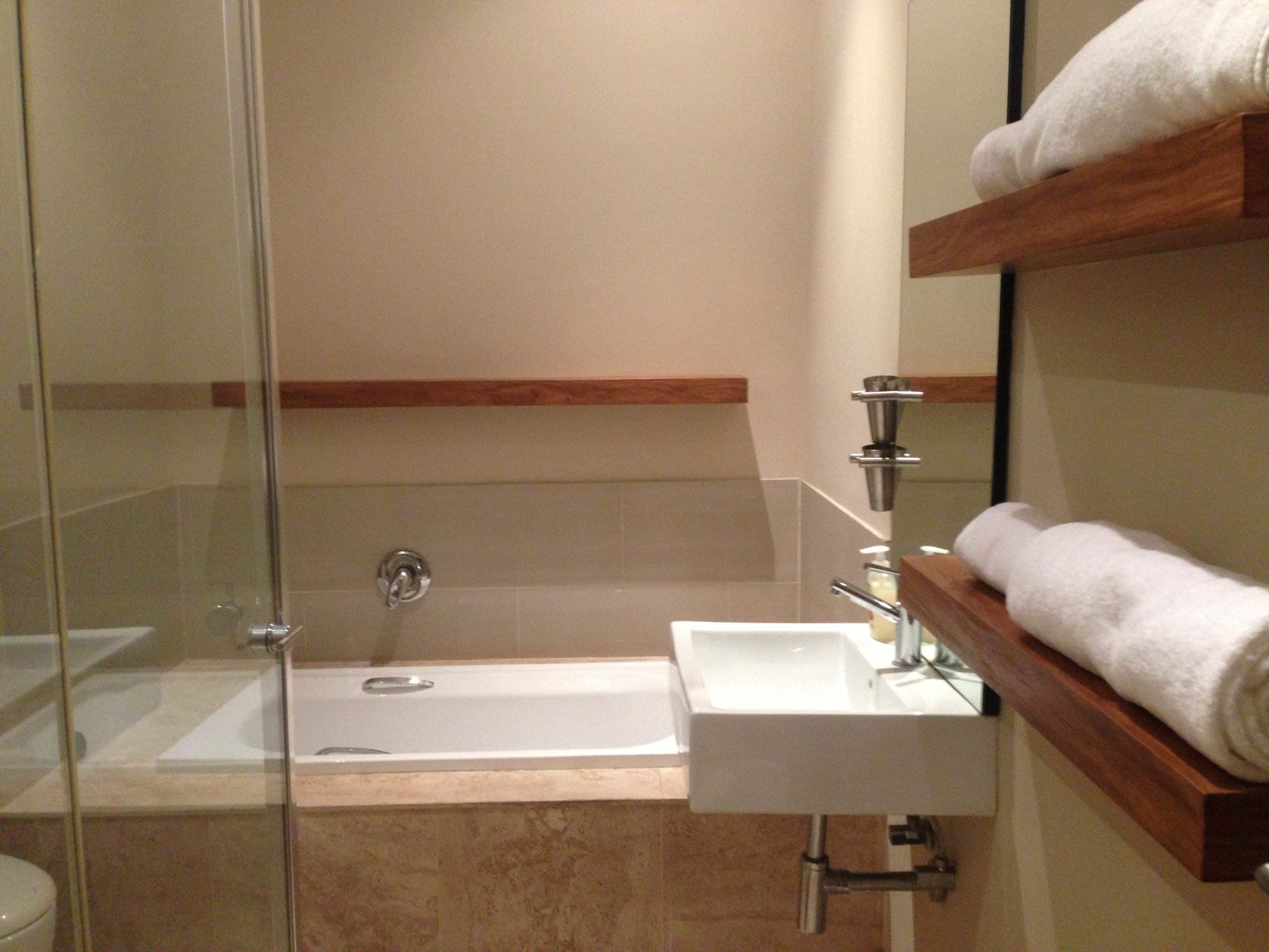 A modern bathroom with a glass shower enclosure, a white sink, and a wooden shelf holding a towel.