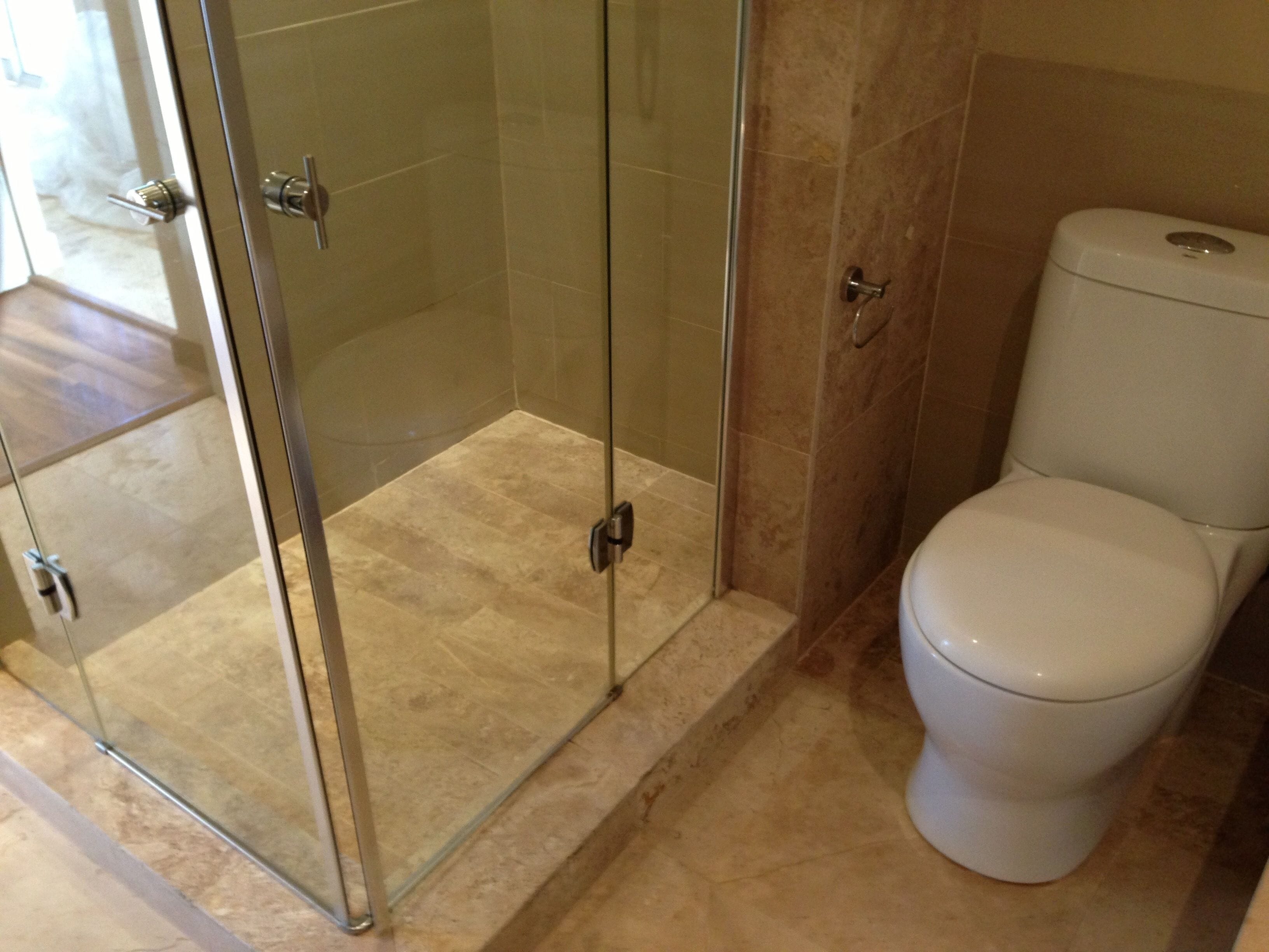 A bathroom with a glass shower enclosure, a toilet, and beige tiled flooring.