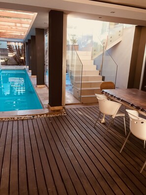 A modern indoor pool area with a wooden deck, a glass staircase, and a dining area with white chairs. - Sunset Beach Guesthouse (Cape Town)
