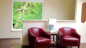 Una habitación con dos sillones de cuero rojo, una mesita con lámpara y un ventanal con vistas al verdor.