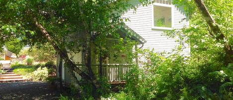 A small, single-story house with a porch, surrounded by lush greenery and a stone pathway leading to the entrance.