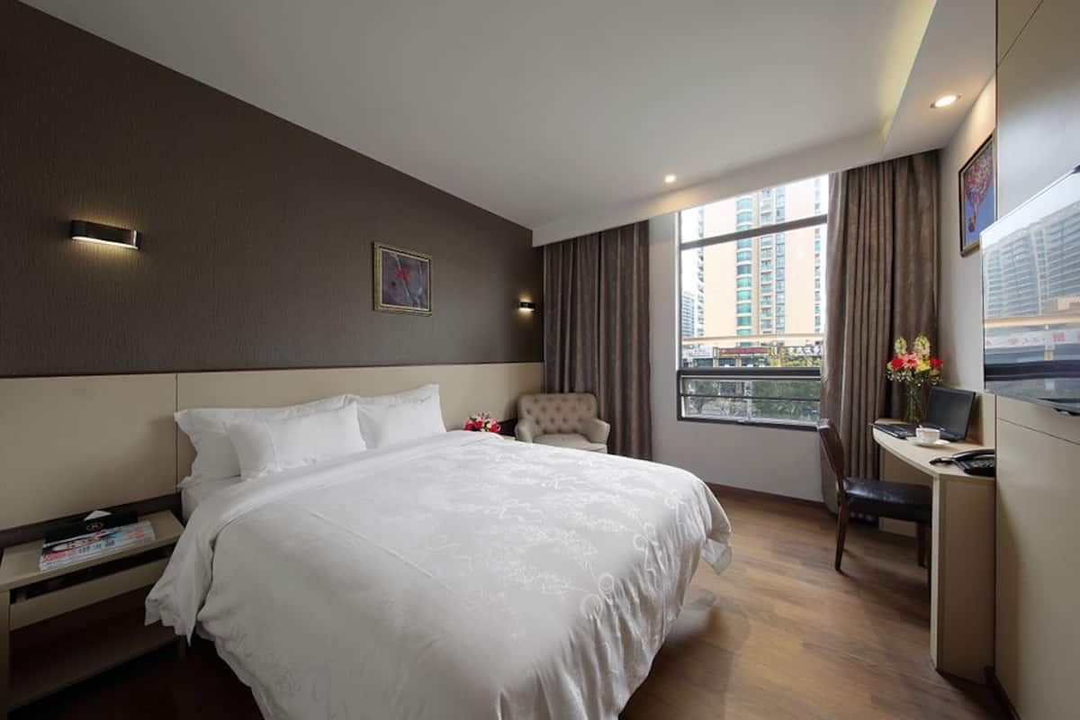 A hotel room with a large bed, a desk with a laptop, a chair, and a view of the city through the window.