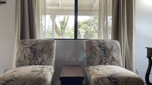 A room with two floral-patterned armchairs facing a window with sheer curtains, a small wooden table, and a wall-mounted sign.