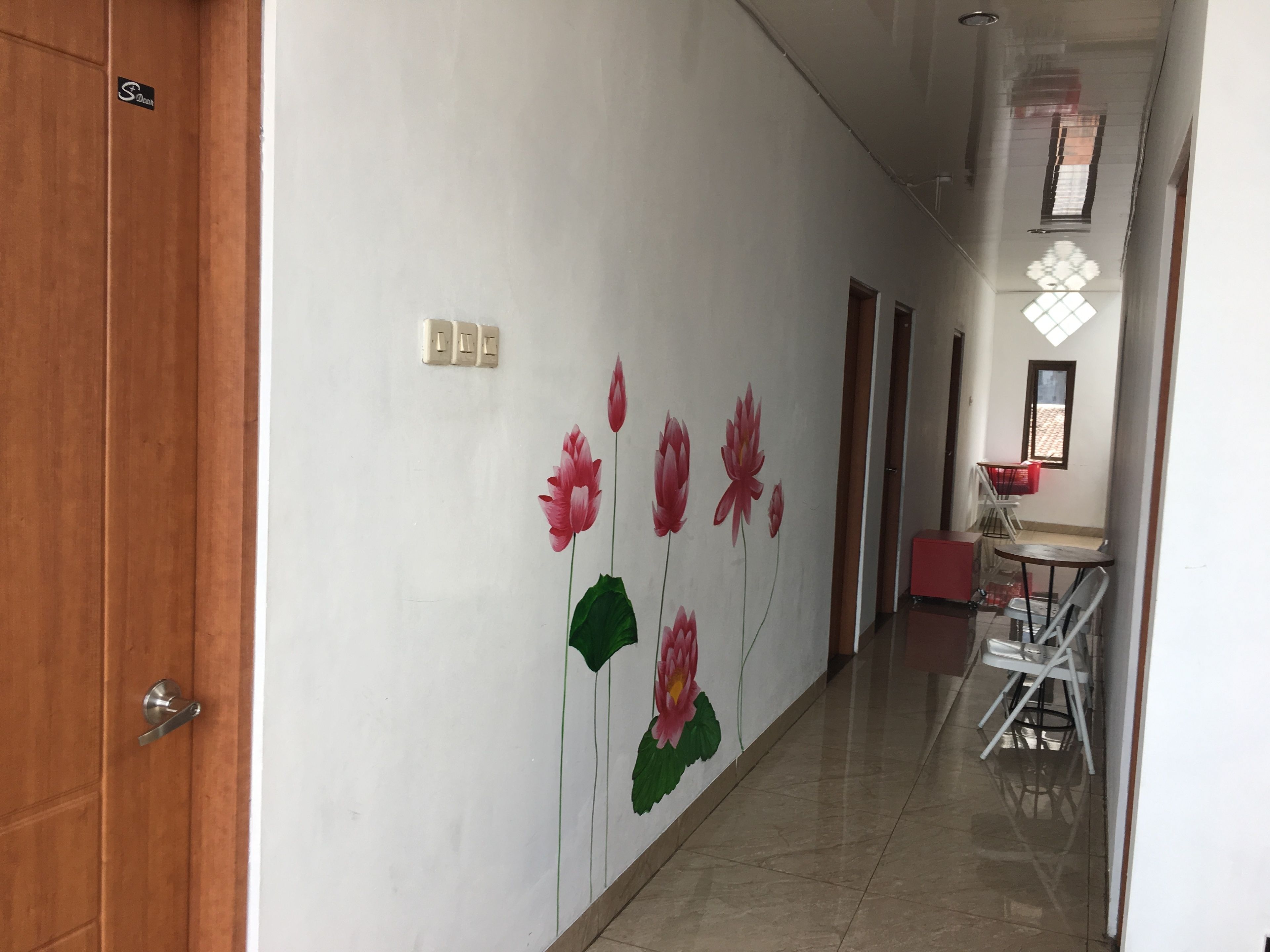 Een hal met een witte muur waarop een muurschildering van een lotusbloem is aangebracht. Links is een houten deur en in de rechterhoek staat een klein tafeltje met stoelen.