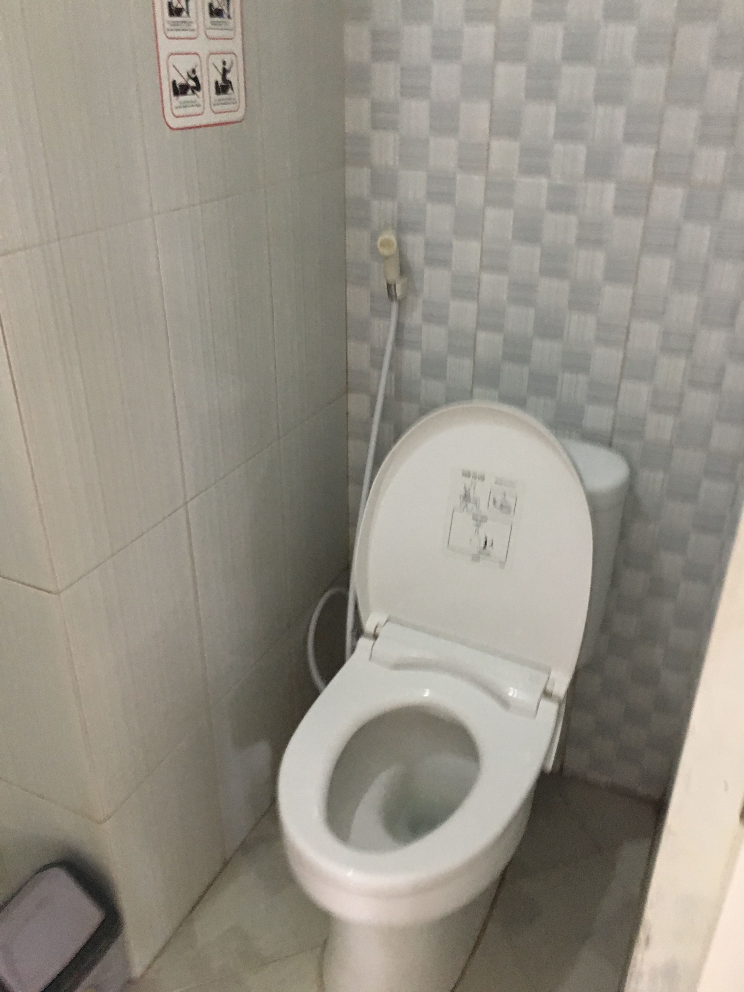 Uma casa de banho pequena, com azulejos, um autoclismo branco, um secador de mãos e um botão de descarga fixado na parede.