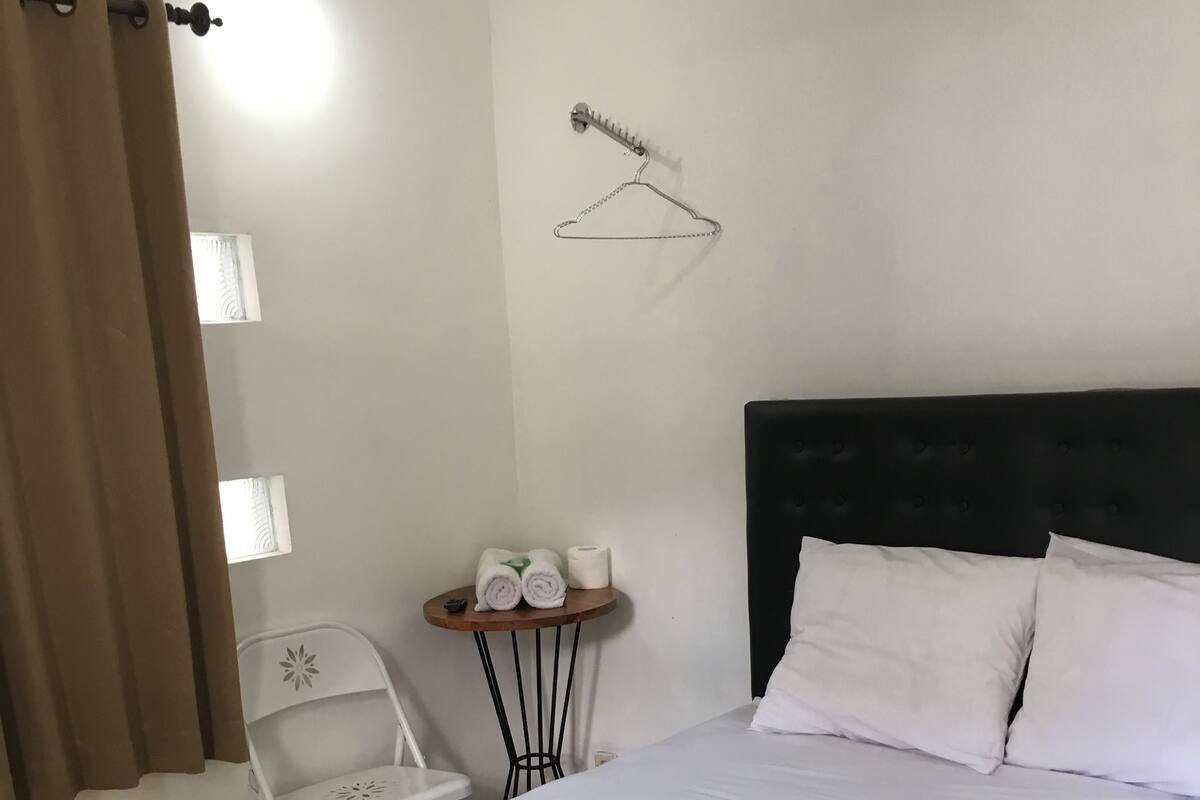 A hotel room with a bed, a small table with towels, a chair, and a wall-mounted hanger.