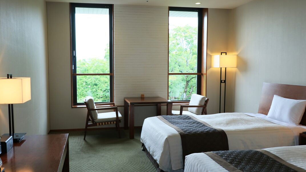 A hotel room with two beds, a desk, a chair, a lamp, and large windows with a view of greenery.
