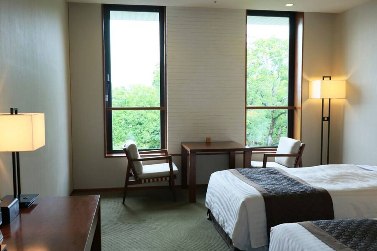 A hotel room with two beds, a desk, a chair, a lamp, and large windows with a view of greenery.