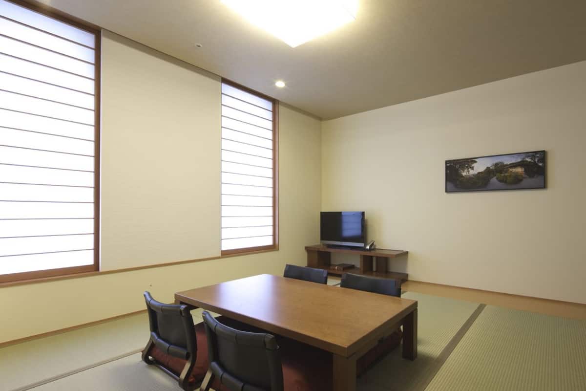 Una habitación con suelo de esteras de tatami, una mesa de madera, sillas, un televisor sobre un pedestal y un cuadro en la pared.