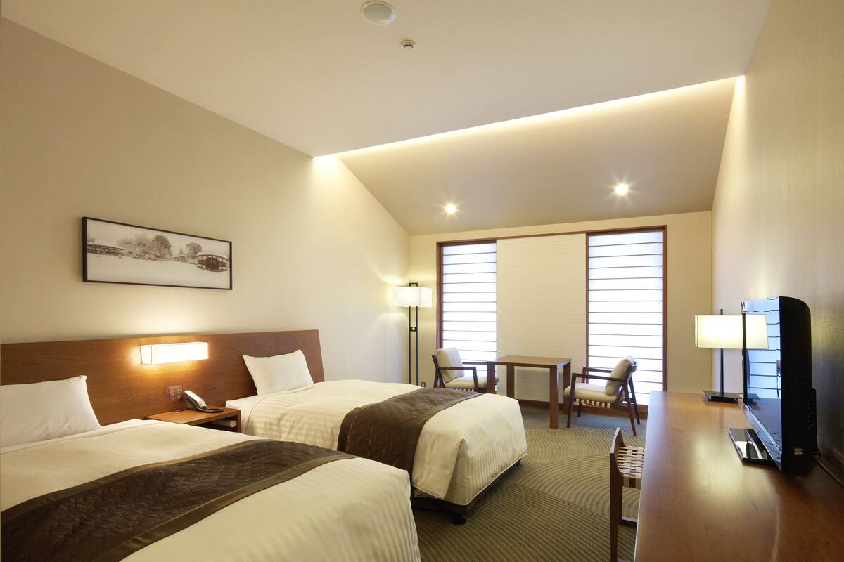 Habitación de hotel con dos camas, un escritorio, una silla, un televisor y una ventana con persianas.