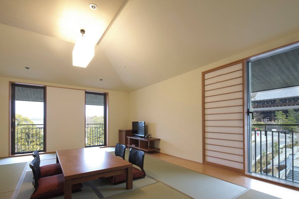 Una habitación con suelo de tatami, una mesa baja, sillas, televisor y ventanas amplias con vistas al exterior.