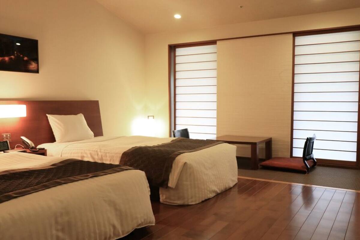 A hotel room with two beds, a desk, a chair, a nightstand, a lamp, and a large window with blinds.