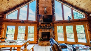 Interior de una cabaña de troncos con chimenea de piedra, techo de madera, ventanas amplias y una terraza de madera afuera.