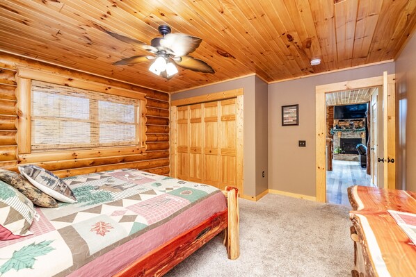 A log cabin bedroom with a bed, wooden walls, a ceiling fan, and a view into the living area.