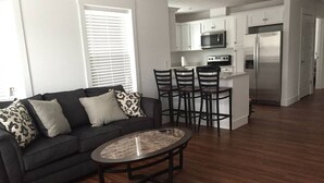 A modern living room with a dark sofa, patterned cushions, a glass coffee table, and a kitchen area with white cabinets, a microwave, and a refrigerator.