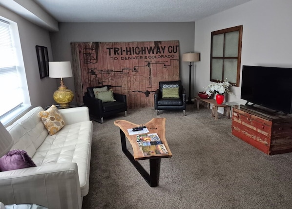 A living room with a white sofa, a black leather chair, a wooden coffee table, a TV stand, and a map mural on the wall.