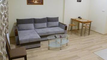 Un salon avec un canapé gris, une table basse en verre, une table à manger en bois avec deux chaises, et un petit tableau accroché au mur.