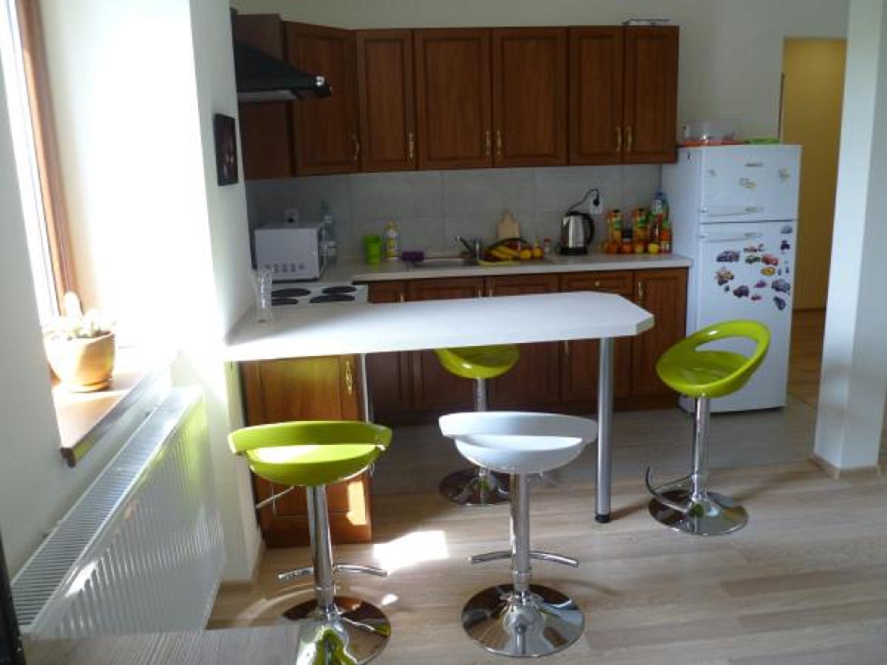 Una cocina con armarios de madera, una encimera blanca y un refrigerador blanco. Hay dos taburetes verdes y una silla blanca.