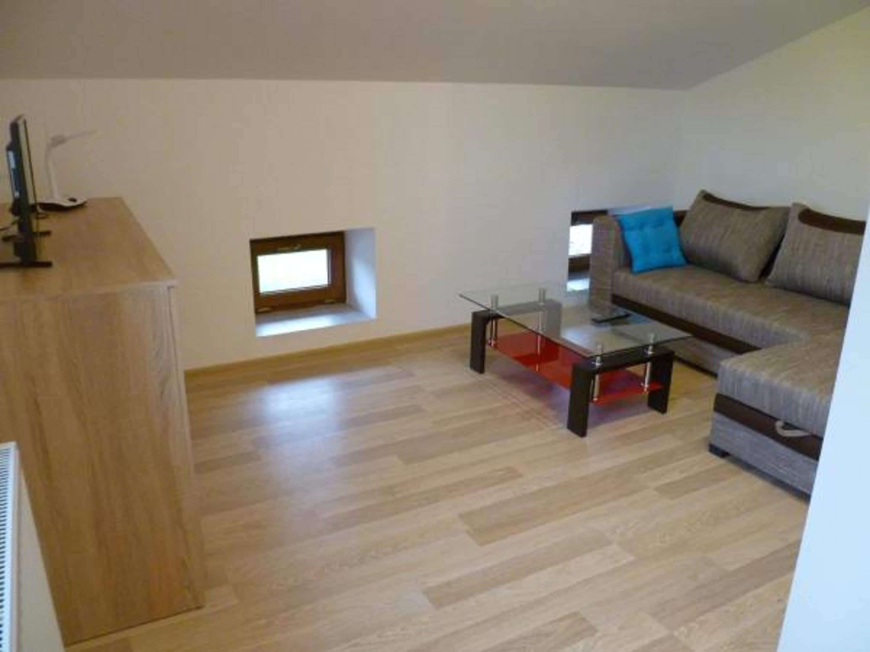 Ein modernes Wohnzimmer mit einem Glastisch, einer Couch mit Kissen, einem kleinen Fenster und einer Holzkommode.