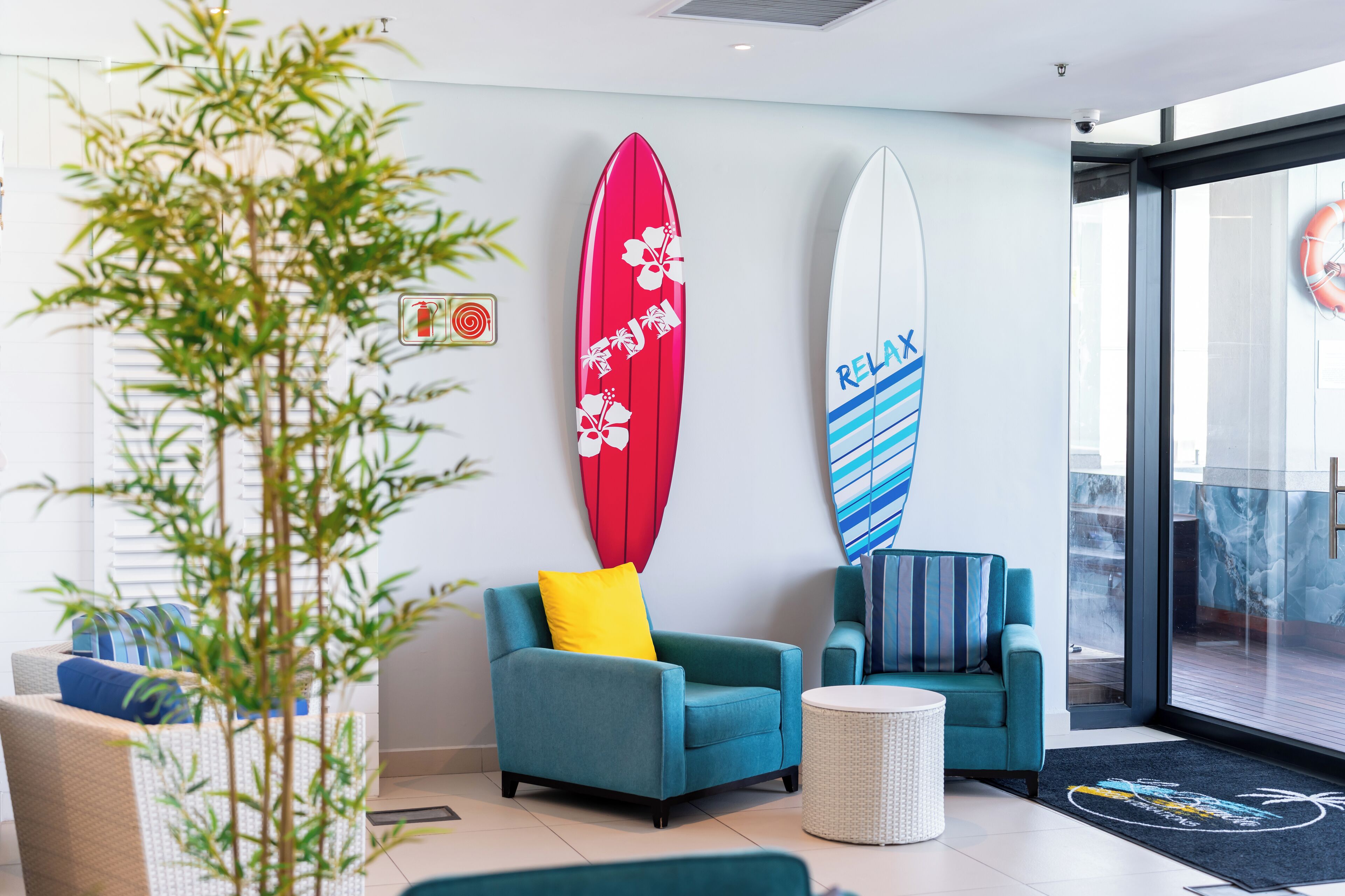 A modern lobby with teal armchairs, a large surfboard decoration, and a glass door leading to an outdoor area.