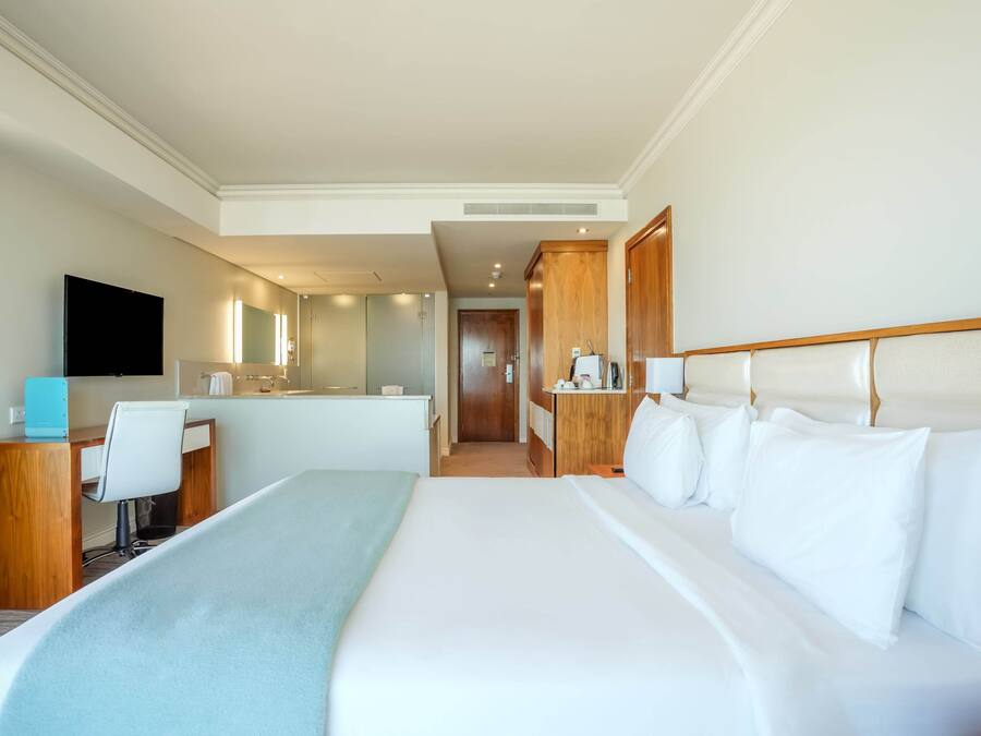 A hotel room with a large bed, a desk, a chair, a TV, and a bathroom visible in the background.