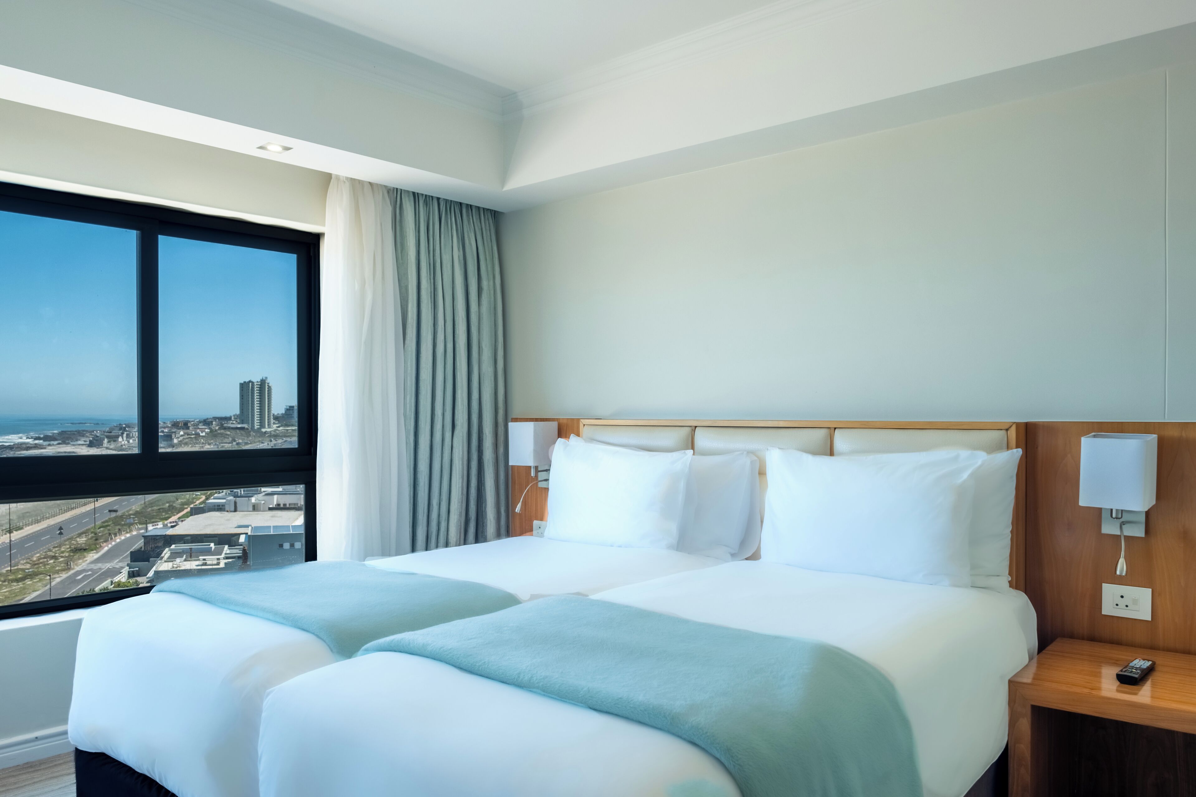 A hotel room with a large bed, a nightstand, a lamp, and a view of the city through a large window.