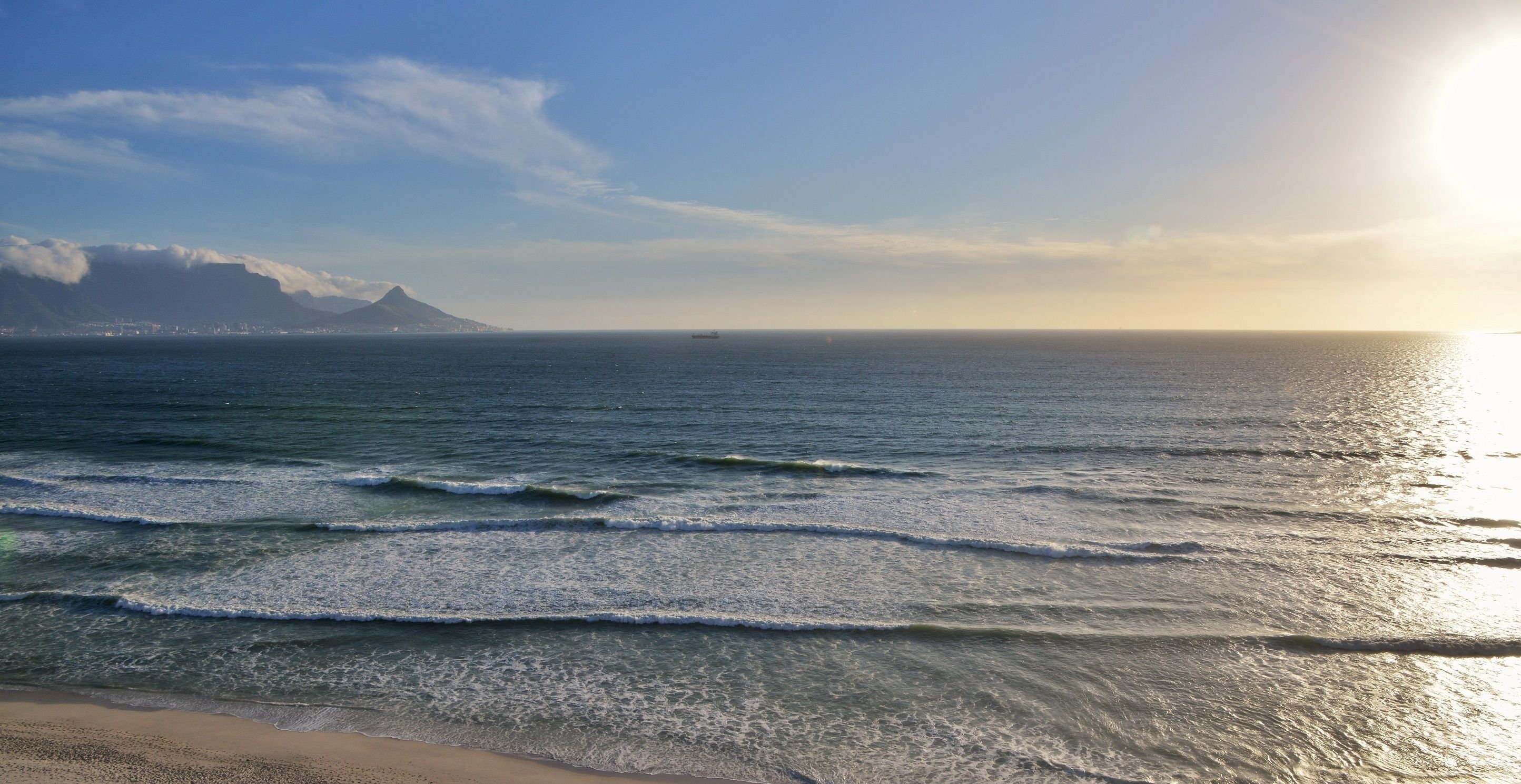 A beach with waves, a distant mountain, and a ship in the distance.