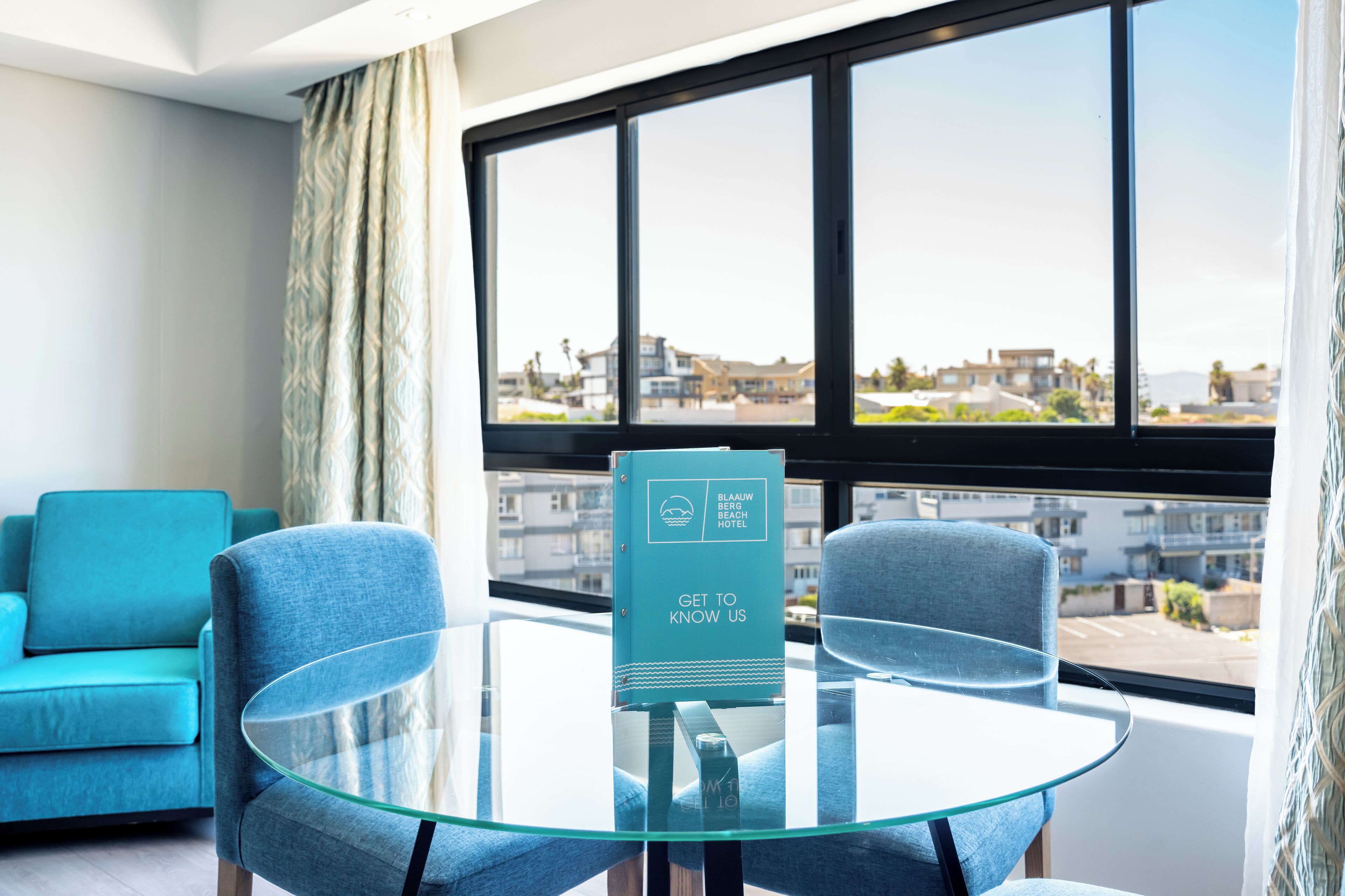 Een moderne hotelkamer met een glazen tafel, twee blauwe fauteuils, uitzicht op de stad door grote ramen en een bordje op de tafel.