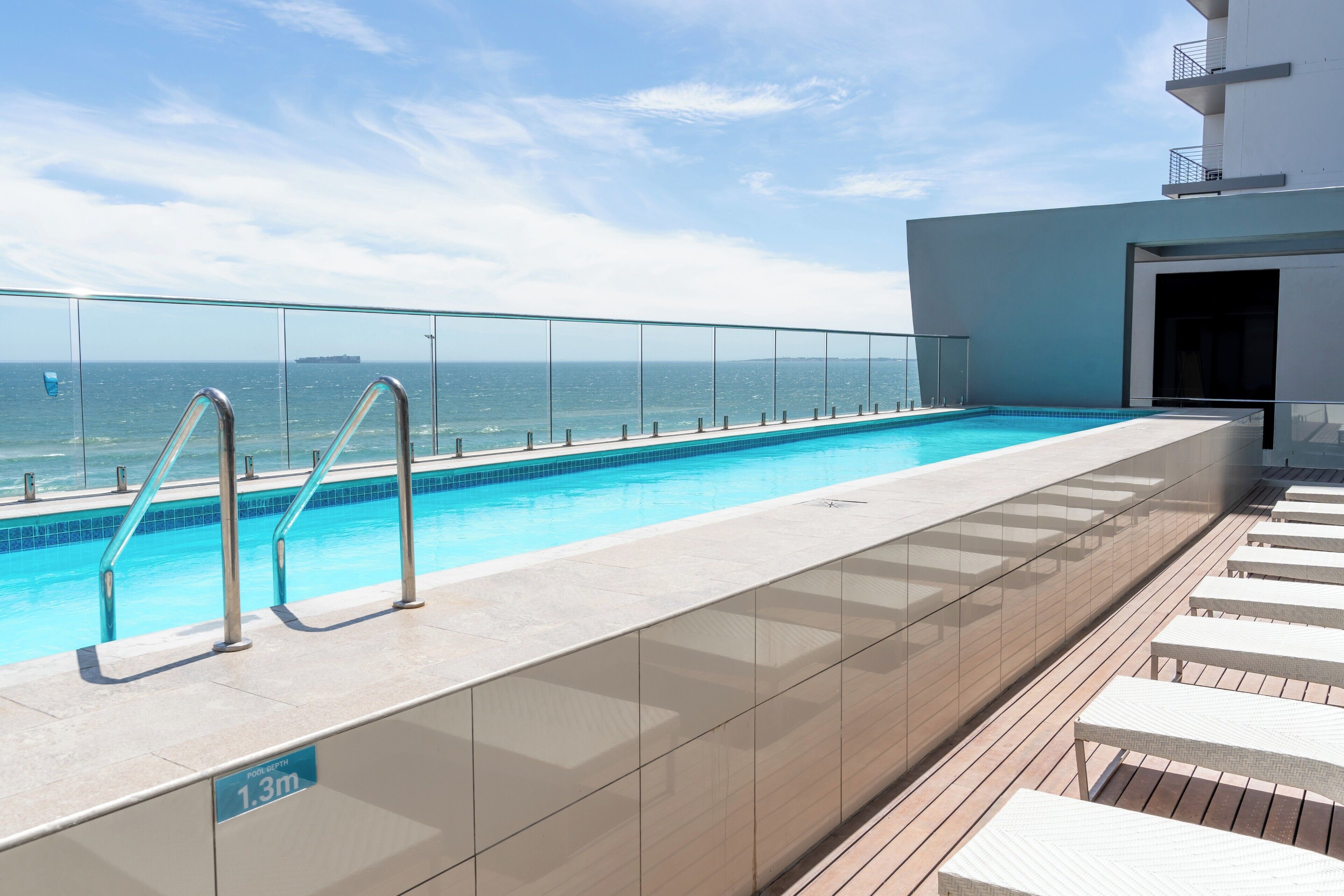 Una piscina en la azotea con barandilla de vidrio, terraza de madera y vista al océano.